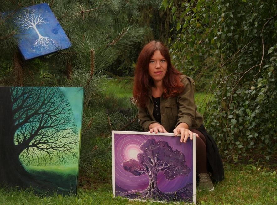 Aleksandra Stencka kuca, przytrzymuje jeden ze swoich obrazów, obok obraz oparty o drzewo, drugi - na drzewie. W tle drzewa.